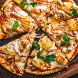 thin crust pizza on a wooden cutting board topped with pineapple, onion, parsley, chicken