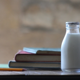 A small bottle of low fat milk next to schoolbooks and a pencil