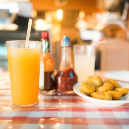 A glass of orange juice with a straw next to a plate of sweet plantains on a checkered tablecloth