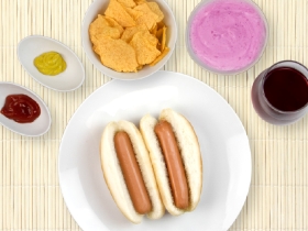 2 hot dogs on a plate surrounded by dips, bowl of chips, blueberry yogurt and cranberry juice