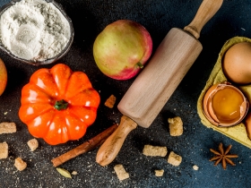 Black background with bowl of flour, small orange pumpkin, apple, rolling pin, carton with one cracked egg, and spices sprinkled in background
