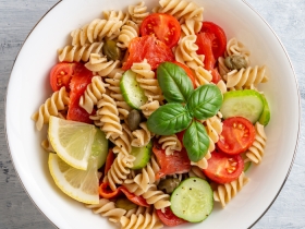 bowl of rotini with cucumber, tomatoes, olives, basil and lemon wedges