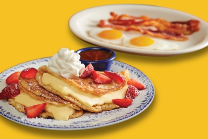 foreground: stuffed cheesecake pancake topped with whipped cream and strawberries on plate. background: two eggs, sunny-side-up and strips of bacon on plate