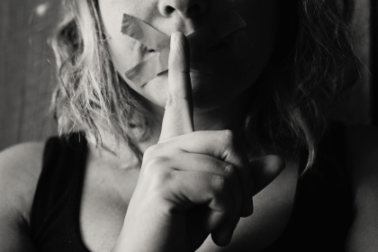 A woman with tape over her mouth making a shush gesture