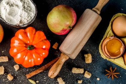 Black background with bowl of flour, small orange pumpkin, apple, rolling pin, carton with one cracked egg, and spices sprinkled in background