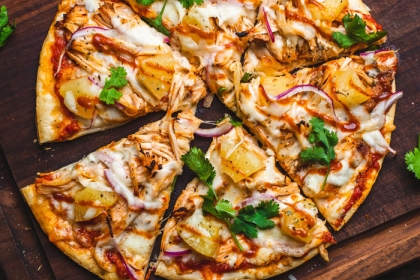 thin crust pizza on a wooden cutting board topped with pineapple, onion, parsley, chicken
