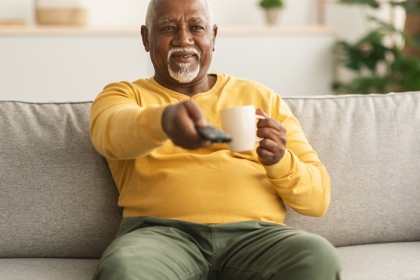 older man holding coffee and pointing remote towards the tv while sitting on a sofa