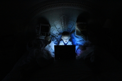 A young boy lit by the glow of an electronic screen. He is sitting between his parents, in bed, but is not being monitored.