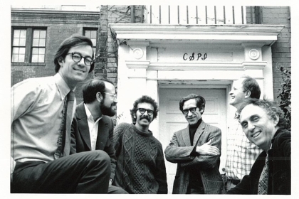 A black and white photograph of five men in front of a building bearing the initials "CSPI"