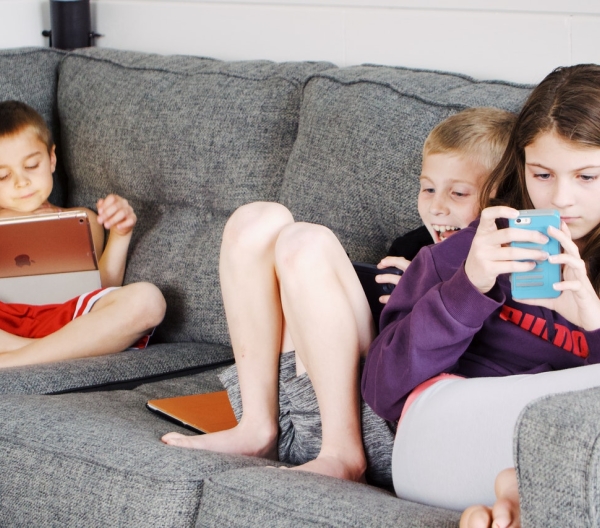 Three young children sit on a couch, looking at phones and tablets