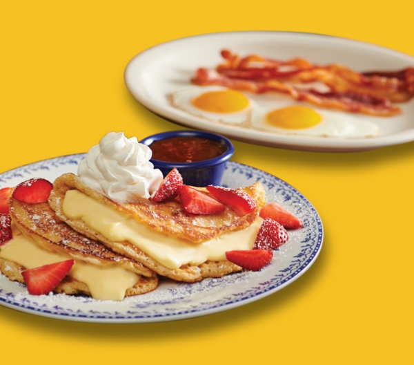 foreground: stuffed cheesecake pancake topped with whipped cream and strawberries on plate. background: two eggs, sunny-side-up and strips of bacon on plate