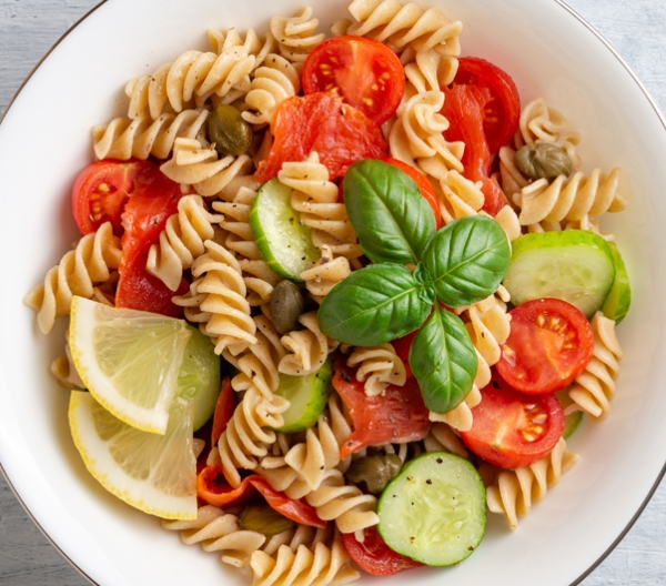 bowl of rotini with cucumber, tomatoes, olives, basil and lemon wedges