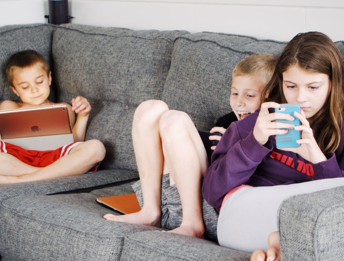 Three young children sit on a couch, looking at phones and tablets