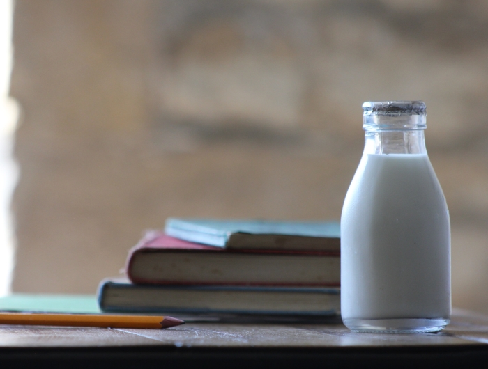 A small bottle of low fat milk next to schoolbooks and a pencil