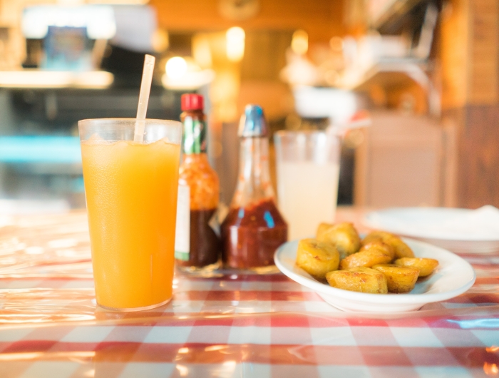 A glass of orange juice with a straw next to a plate of sweet plantains on a checkered tablecloth