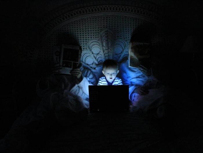A young boy lit by the glow of an electronic screen. He is sitting between his parents, in bed, but is not being monitored.