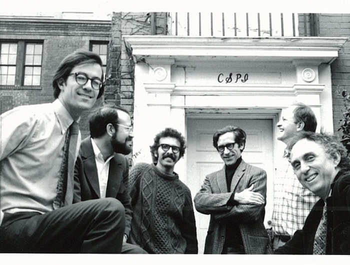 A black and white photograph of five men in front of a building bearing the initials "CSPI"