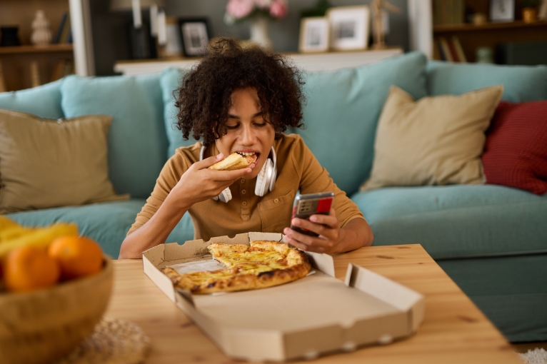 A teen eats pizza at home while using phone