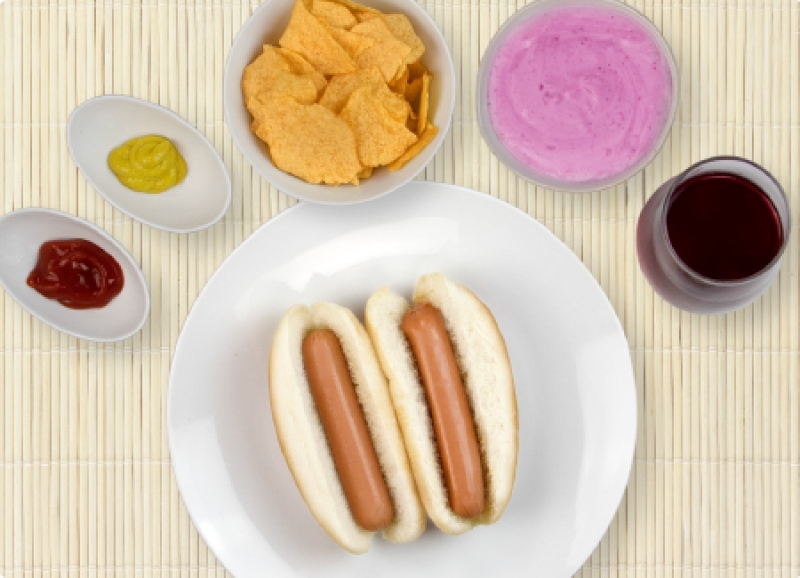2 hot dogs on a plate surrounded by condiments, bowl of chips, yogurt, and cranberry juice drink
